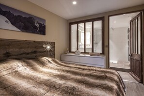 Un dormitorio con una cama grande, cabecera de madera, un ventanal amplio, un baño visible a través del ventanal y una puerta que da a una zona de ducha.