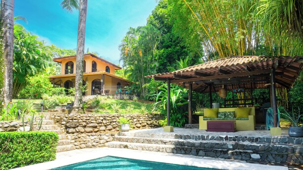 A pool area with a stone wall, a covered seating area, and a building with a tiled roof. - Casa Paraiso - Tropical Private Pool Near Beach (Esterillos Oeste)