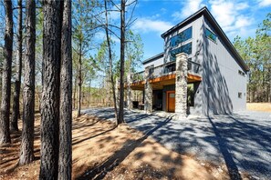 A modern two-story cabin with a stone foundation, large windows, and a balcony overlooking a forested area. - Amazing Outdoor Area:  Pool Table, Hot Tub, Close to Attractions!  (Broken Bow)