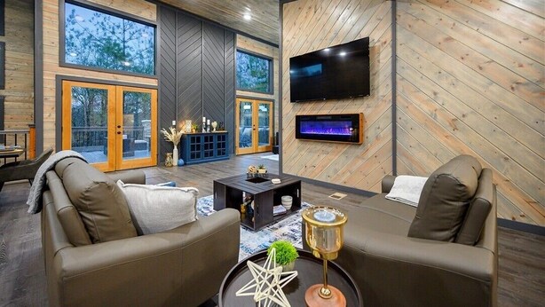 A modern living room with a fireplace, large windows, and a flat-screen TV. - Amazing Outdoor Area:  Pool Table, Hot Tub, Close to Attractions!  (Broken Bow)
