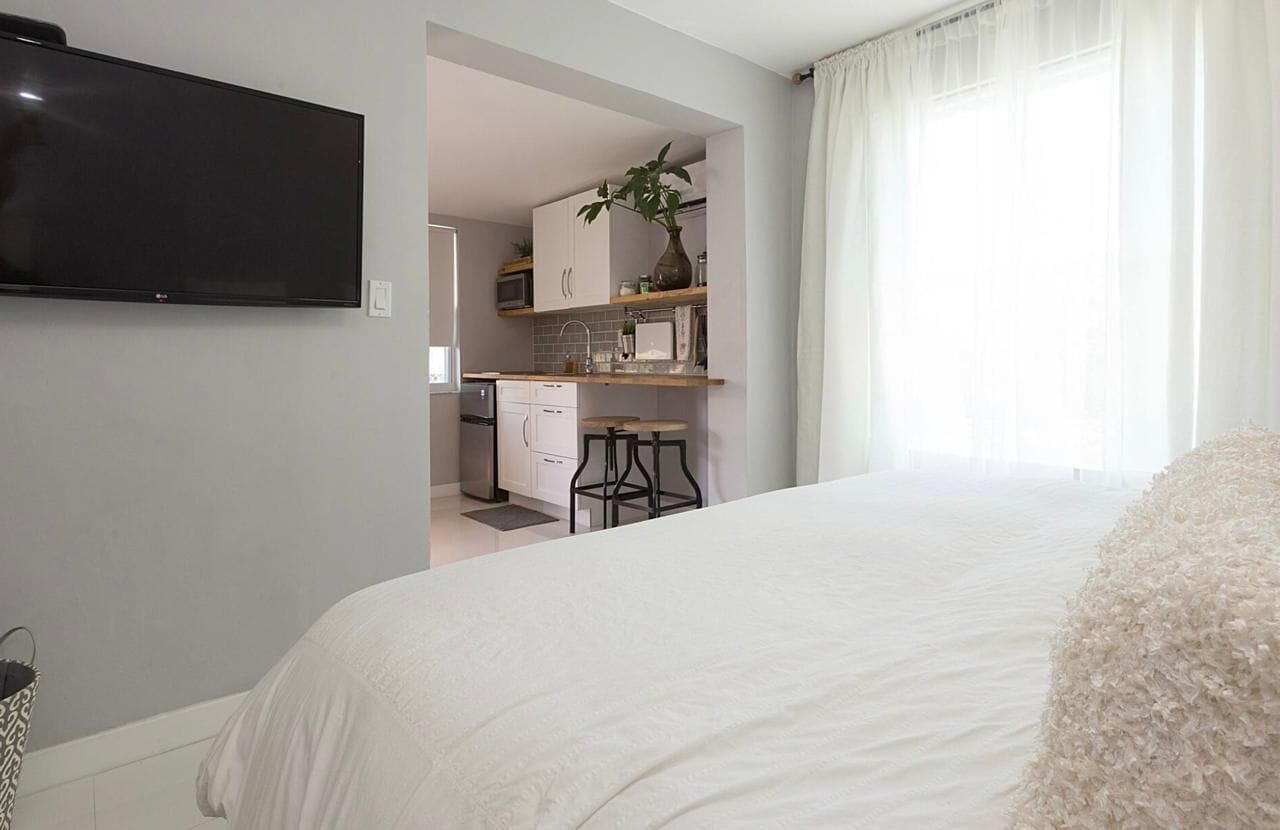 Une chambre avec un grand lit, un téléviseur fixé au mur et une cuisine visible à travers une porte ouverte.