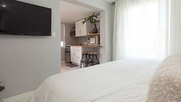 Une chambre avec un grand lit, un téléviseur fixé au mur et une cuisine visible à travers une porte ouverte.