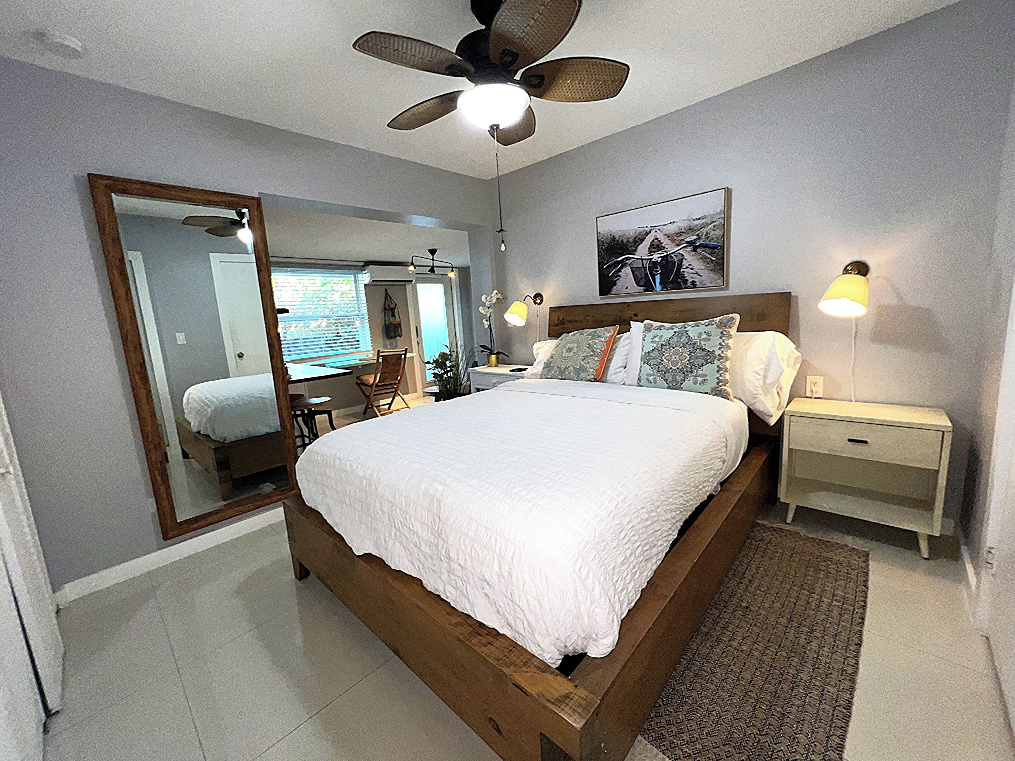 A bedroom with a large bed, a ceiling fan, a bedside table with a lamp, a mirror, and a window with blinds.