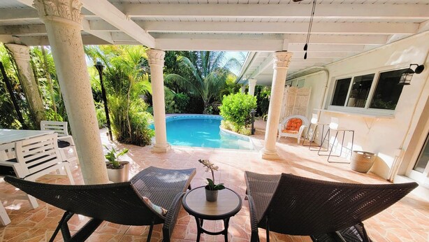 A covered patio with wicker furniture, a pool, and lush greenery.