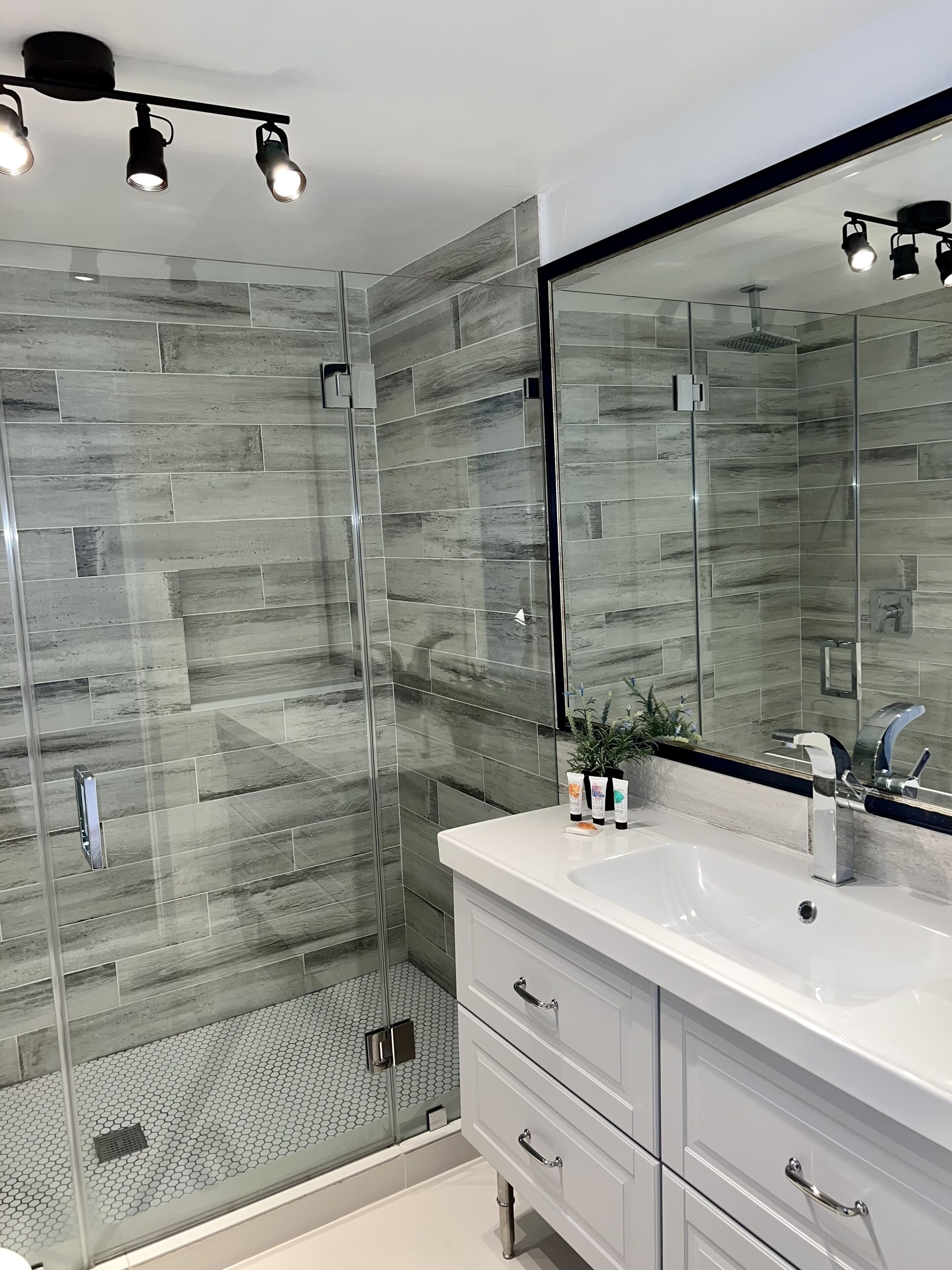 Une salle de bain moderne avec une douche à l’italienne en verre, un meuble-lavabo blanc doté d’un grand miroir et un sol carrelé.