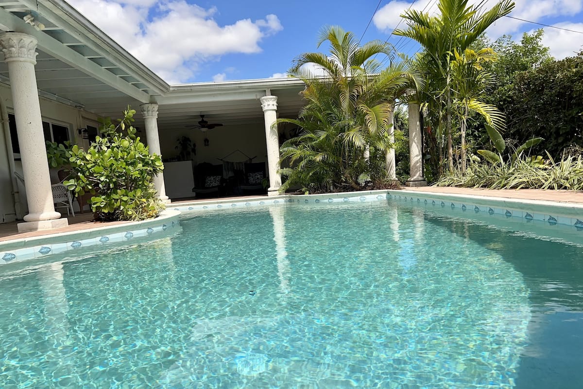 A clear blue pool with a tiled edge, surrounded by lush greenery and a covered patio area with columns.