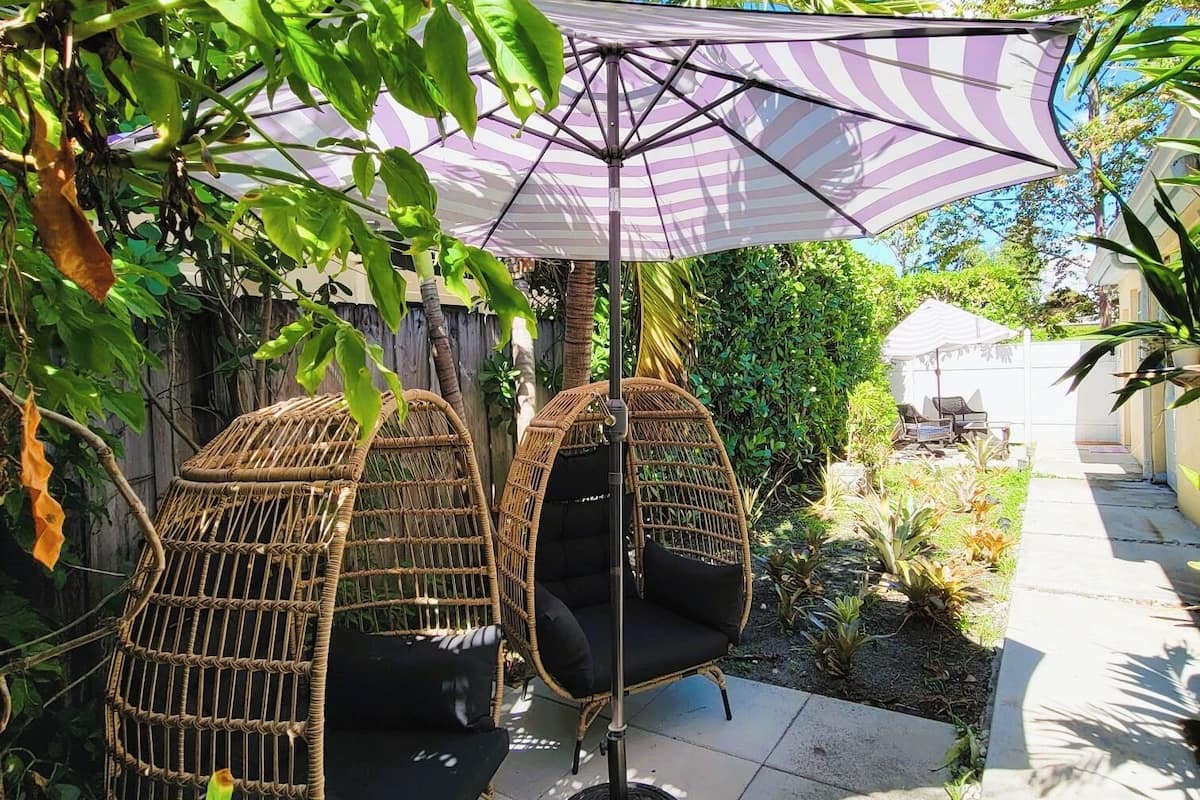 Two wicker swing chairs under a striped umbrella, surrounded by lush greenery and a paved pathway.