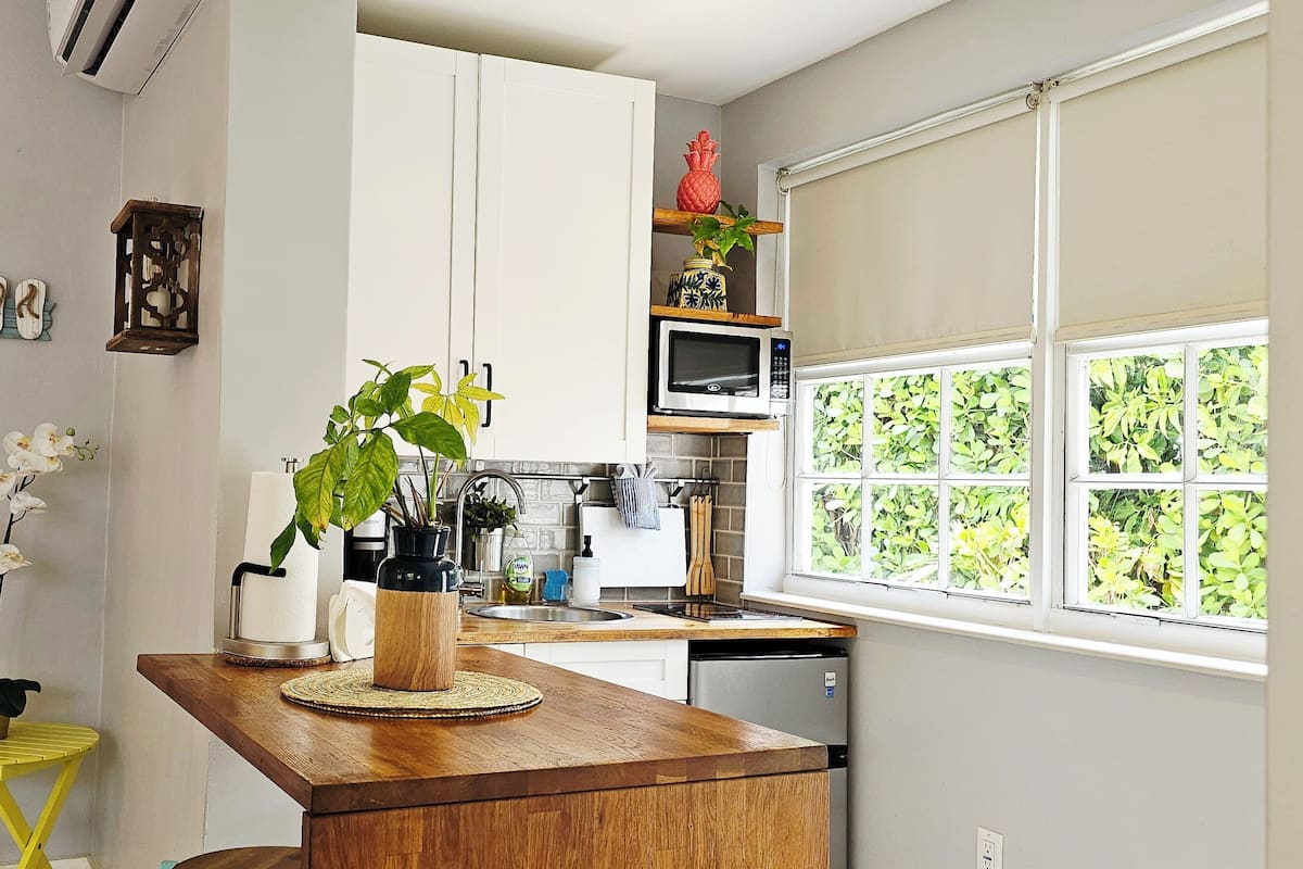 A compact kitchen with white cabinets, a wooden countertop, a small yellow stool, a potted plant, and a window with a view of greenery.