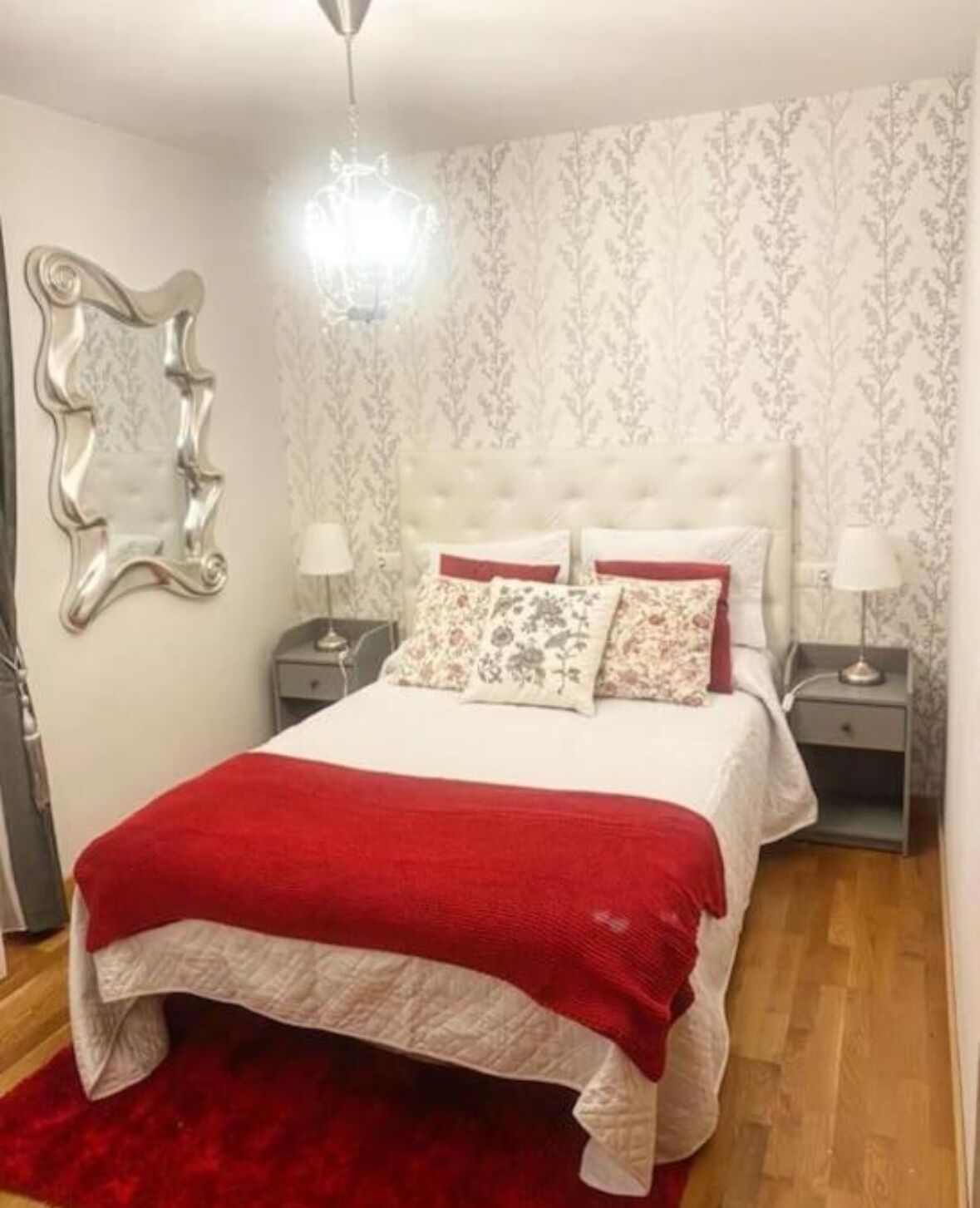 Un dormitorio con cabecera capitonada, una manta roja, almohadas estampadas y dos mesitas de noche con lámparas.