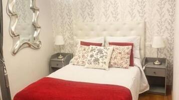 Un dormitorio con cabecera capitonada, una manta roja, almohadas estampadas y dos mesitas de noche con lámparas.
