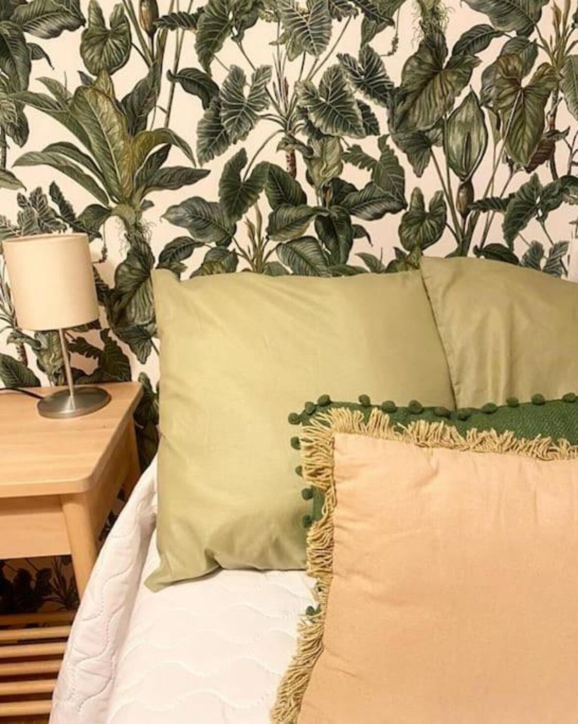Un dormitorio con una cama, almohadas verdes y beige, una mesita de noche de madera y una lámpara.