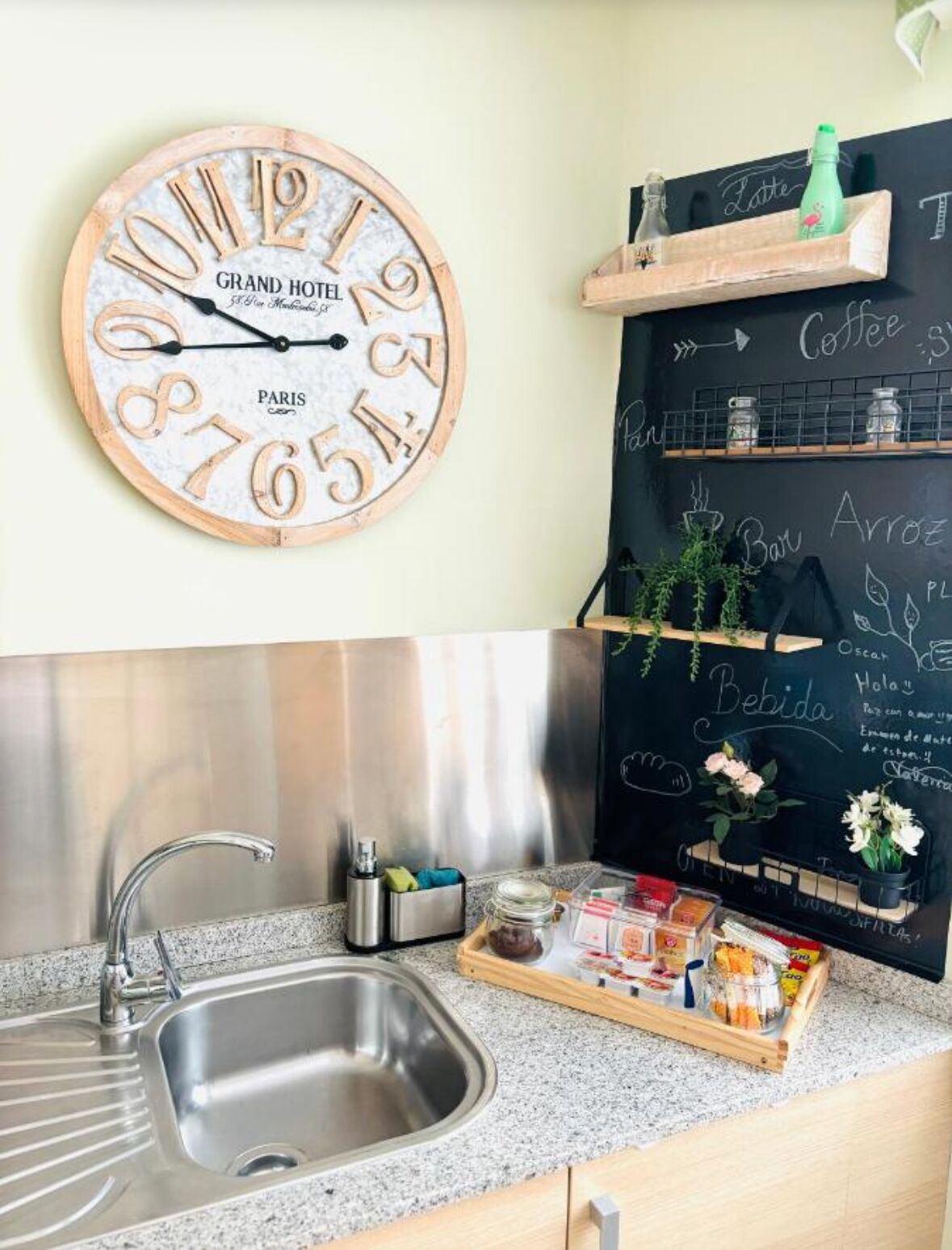A kitchen with a stainless steel sink, a wooden clock with 'Grand Hotel Paris' on it, and a chalkboard menu above the counter.