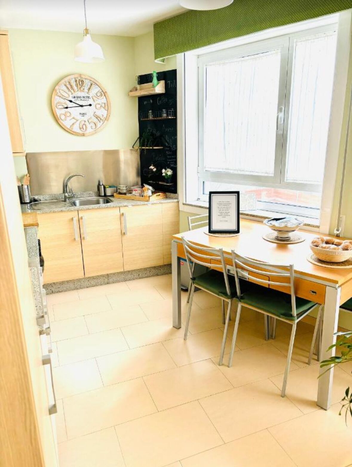 A kitchen with a round table, chairs, a clock, a window, and a chalkboard.