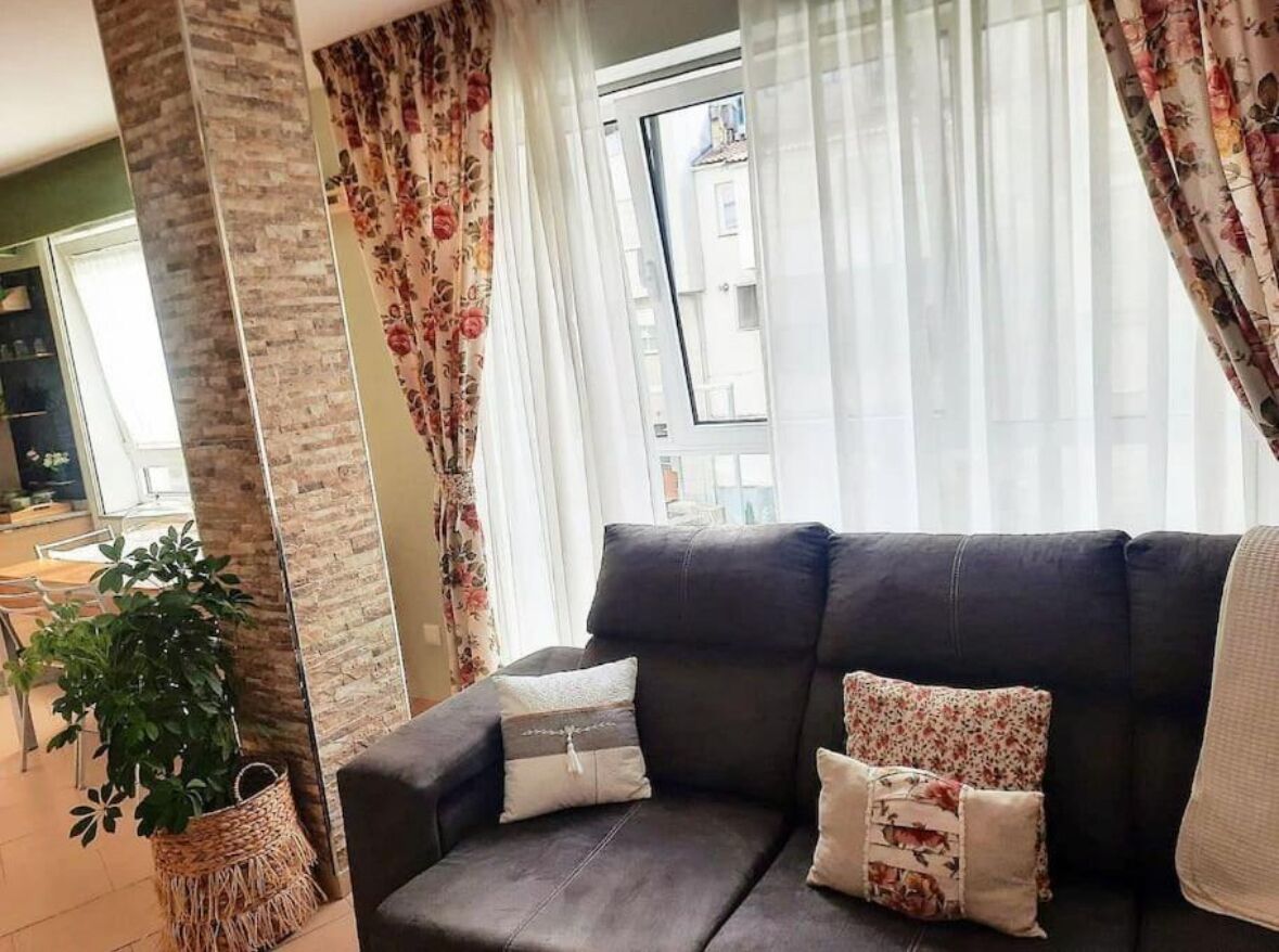 A living room with a dark sofa, floral curtains, a potted plant, and a view of a building through the window.