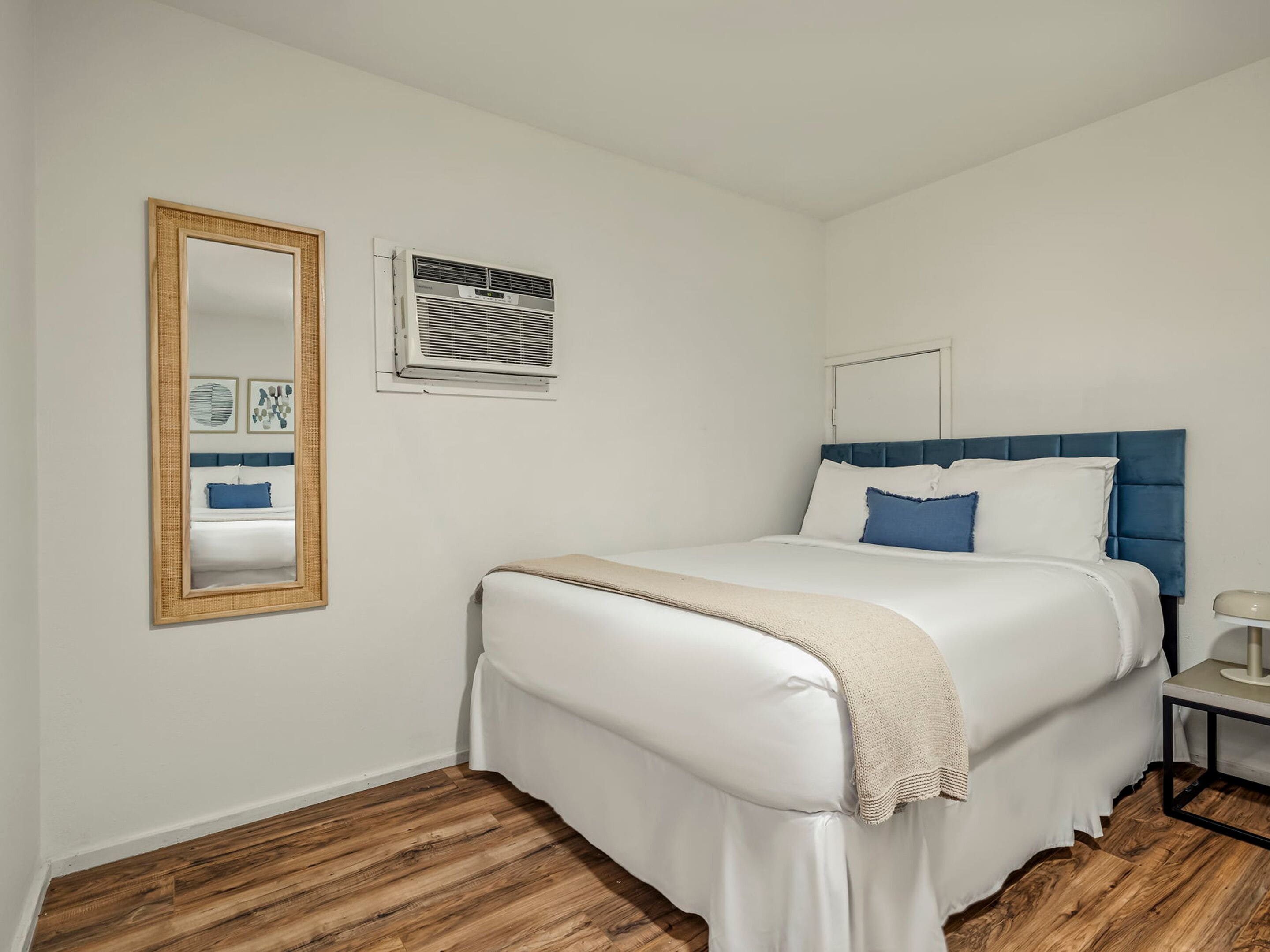 A bedroom with a bed, a bedside table, a mirror, and an air conditioning unit.