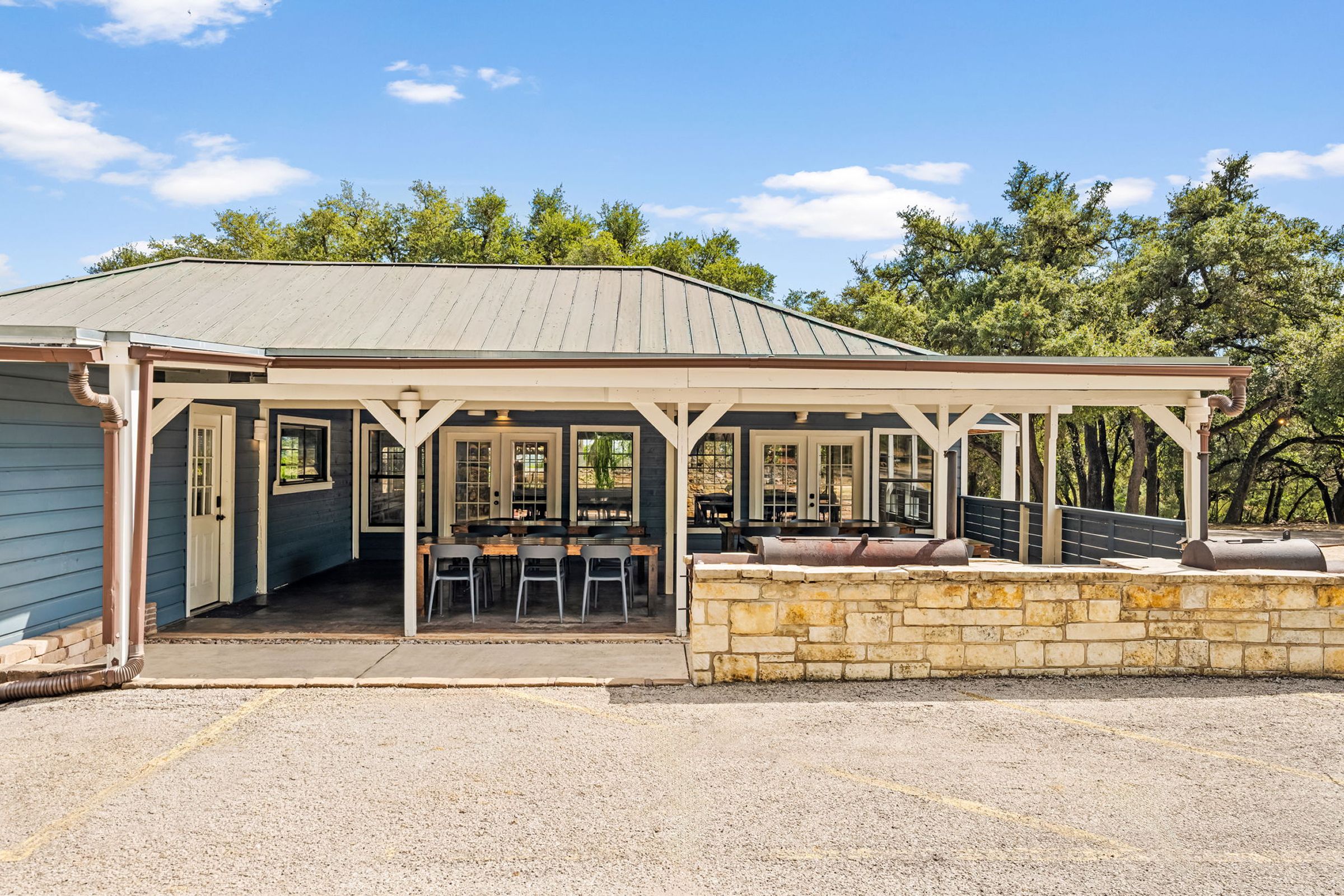 A covered outdoor seating area with tables and chairs, a stone wall, and a building with a sloped roof.