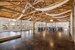 A spacious hall with high ceilings, wooden beams, and large windows. Tables and chairs are arranged for events.