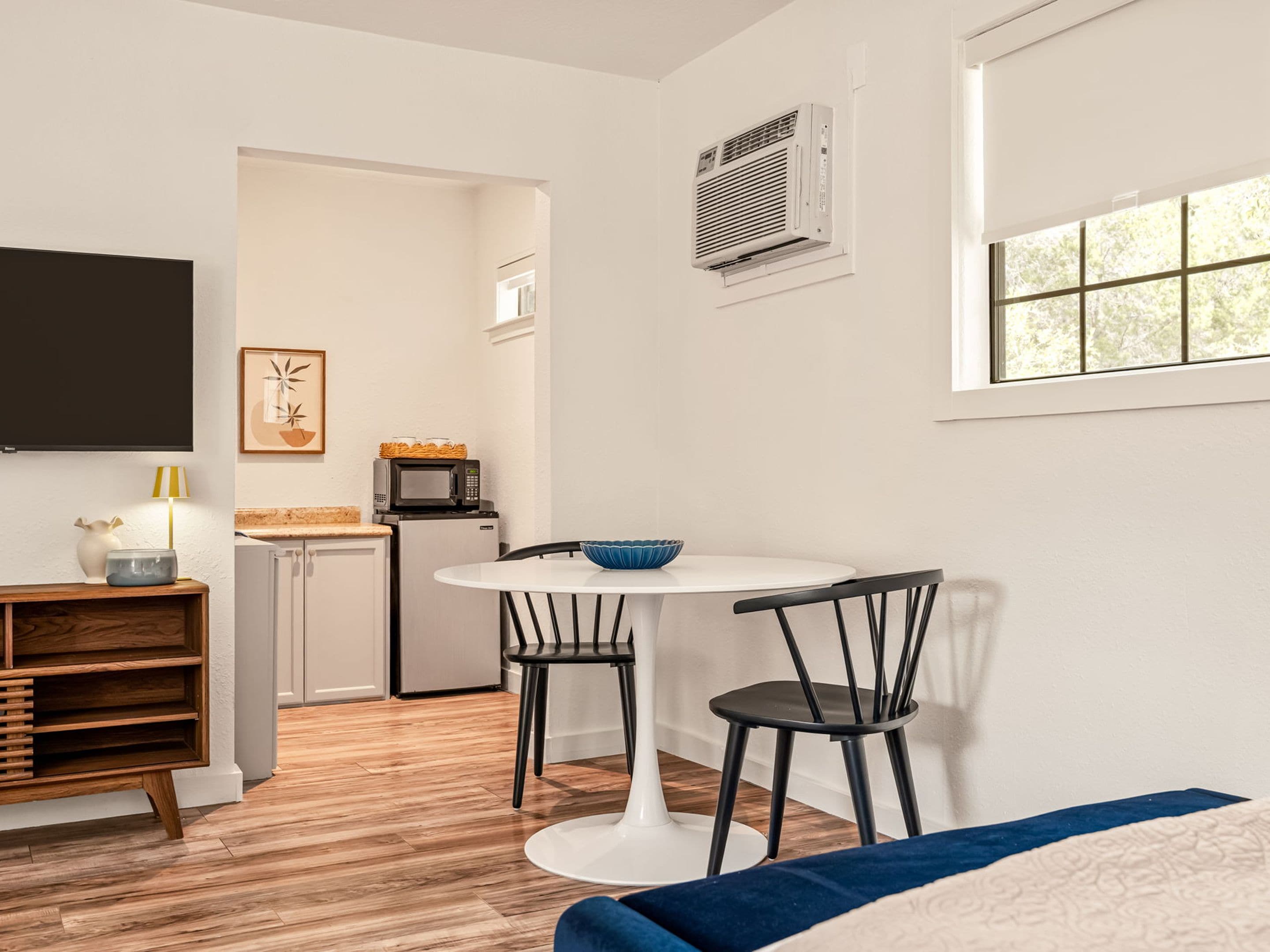 Una habitación pequeña con cama, una mesa de comedor con sillas, microondas, refrigerador y televisor montado en la pared.