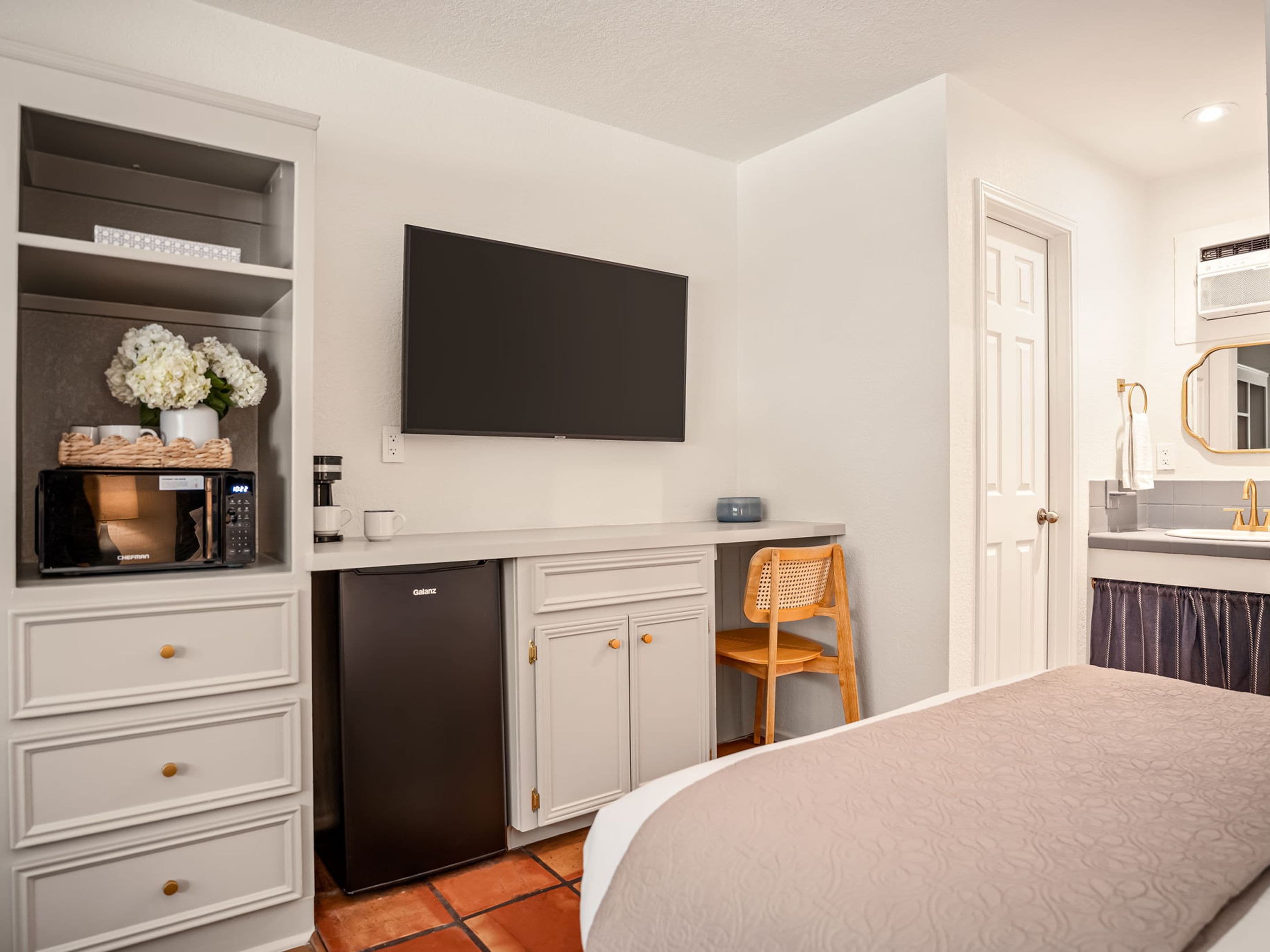 A compact hotel room with a kitchenette, a bed, a flat-screen TV, and a bathroom with a mirror and sink.