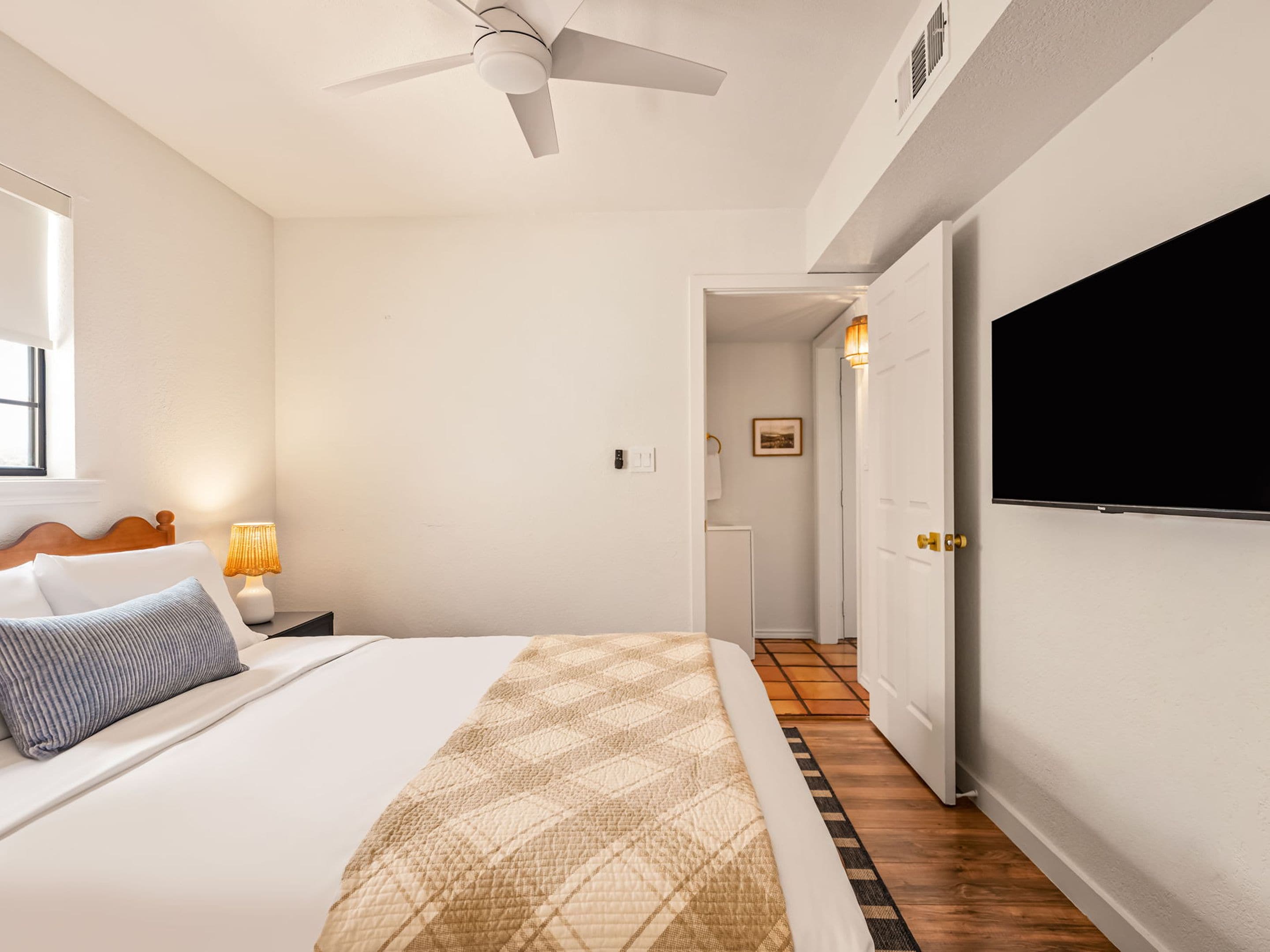A bedroom with a bed, a ceiling fan, a bedside lamp, a TV mounted on the wall, and a door leading to another room.