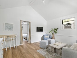 Una habitación moderna y bien iluminada, con suelo de madera, una zona de comedor con taburetes blancos, un espacio de descanso con una silla azul y otomanas, y un gran ventanal con persianas.
