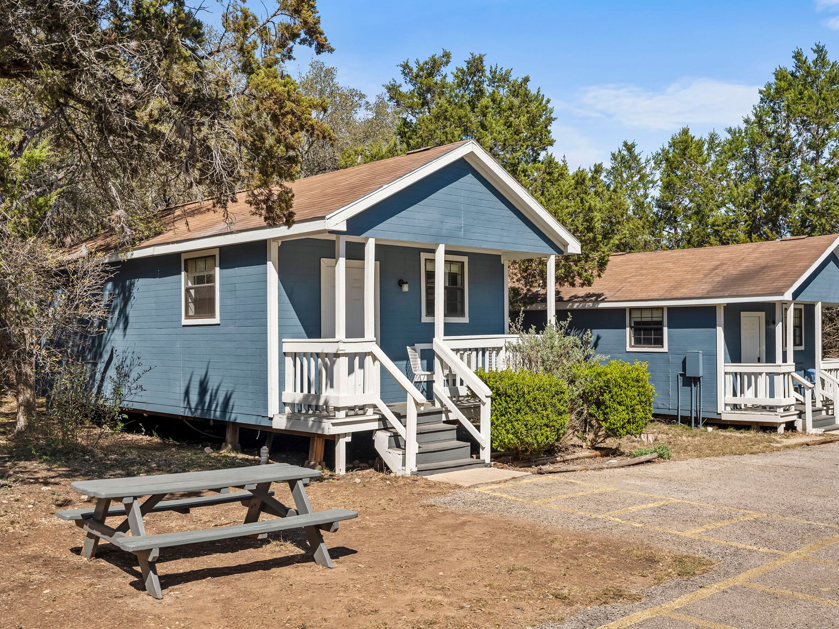 A small, single-story cabin with a porch, a picnic table, and a clear view of the surrounding forest.