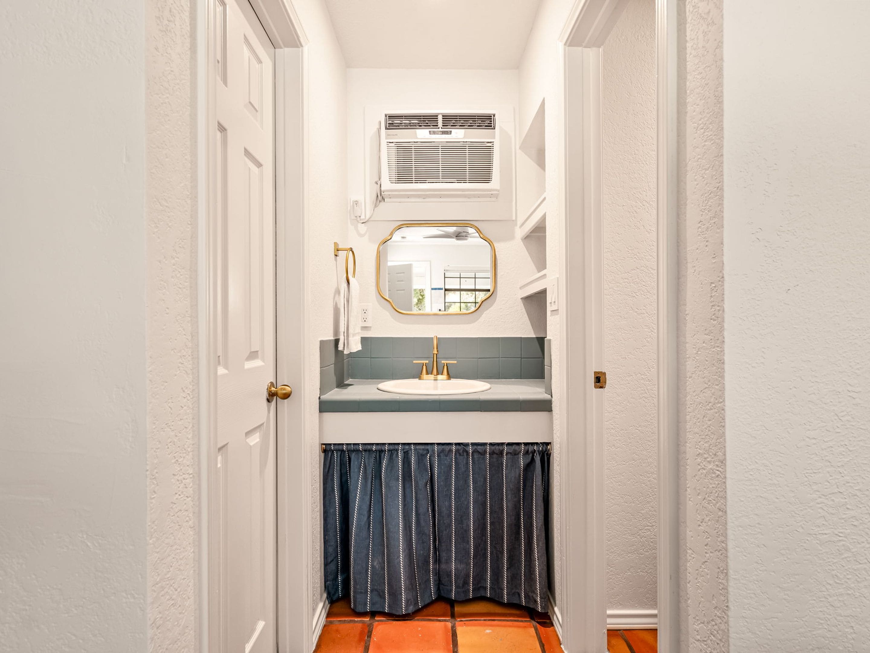 Un pasillo estrecho que conduce a un baño con una puerta blanca, un lavabo blanco, un espejo y un perchero.