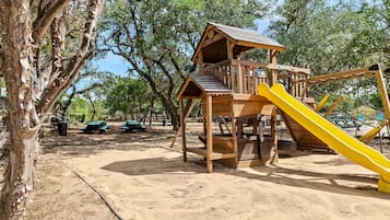 Estructura de madera para juegos con tobogán y zona de escalada, rodeada de árboles y mesas de picnic.