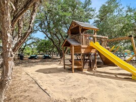 Estructura de madera para juegos con tobogán y zona de escalada, rodeada de árboles y mesas de picnic.