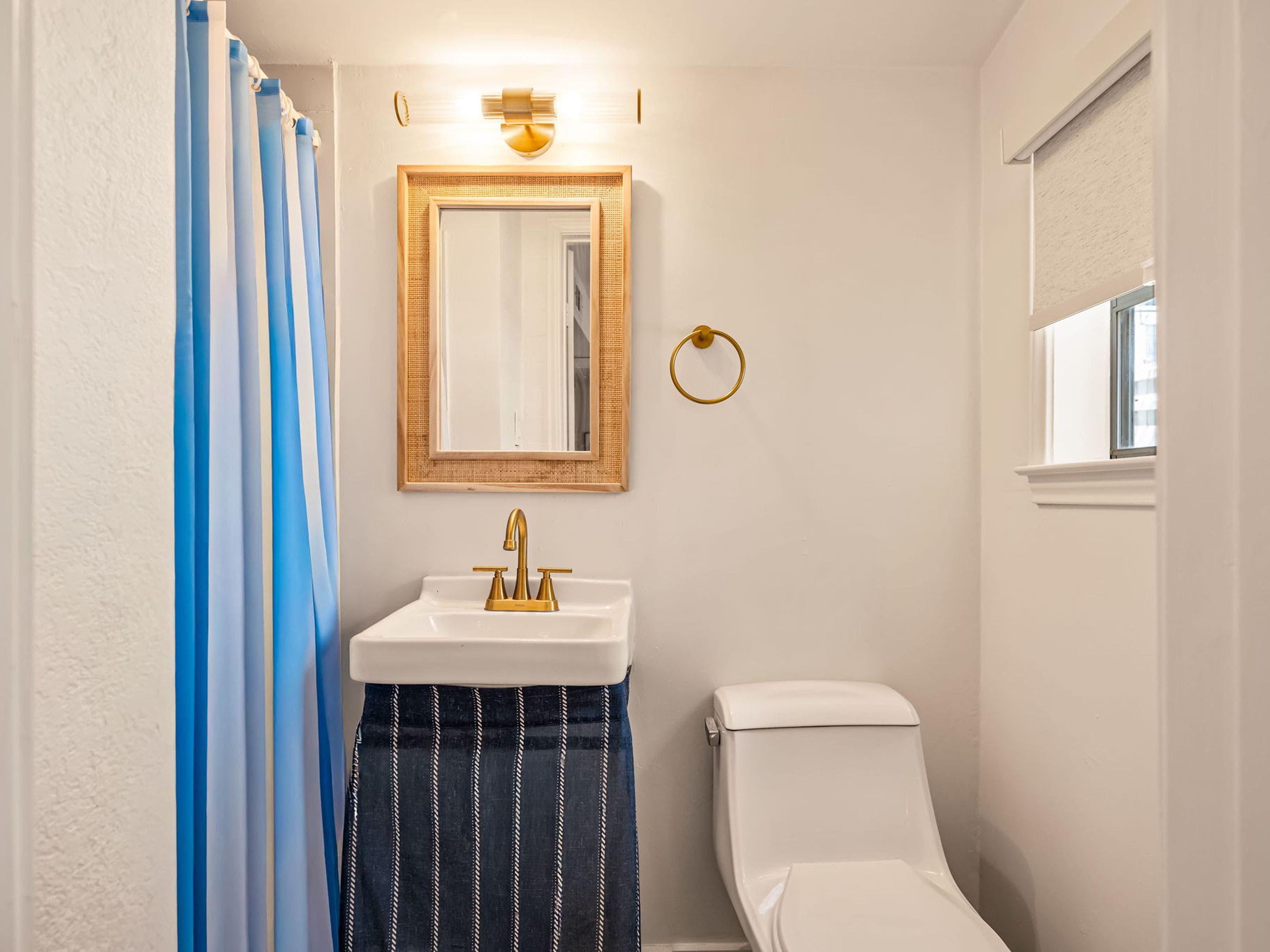 Een badkamer met een witte wastafel, een gouden kraan, een spiegel met een gouden lijst, een toilet en een raam met een jaloezie.