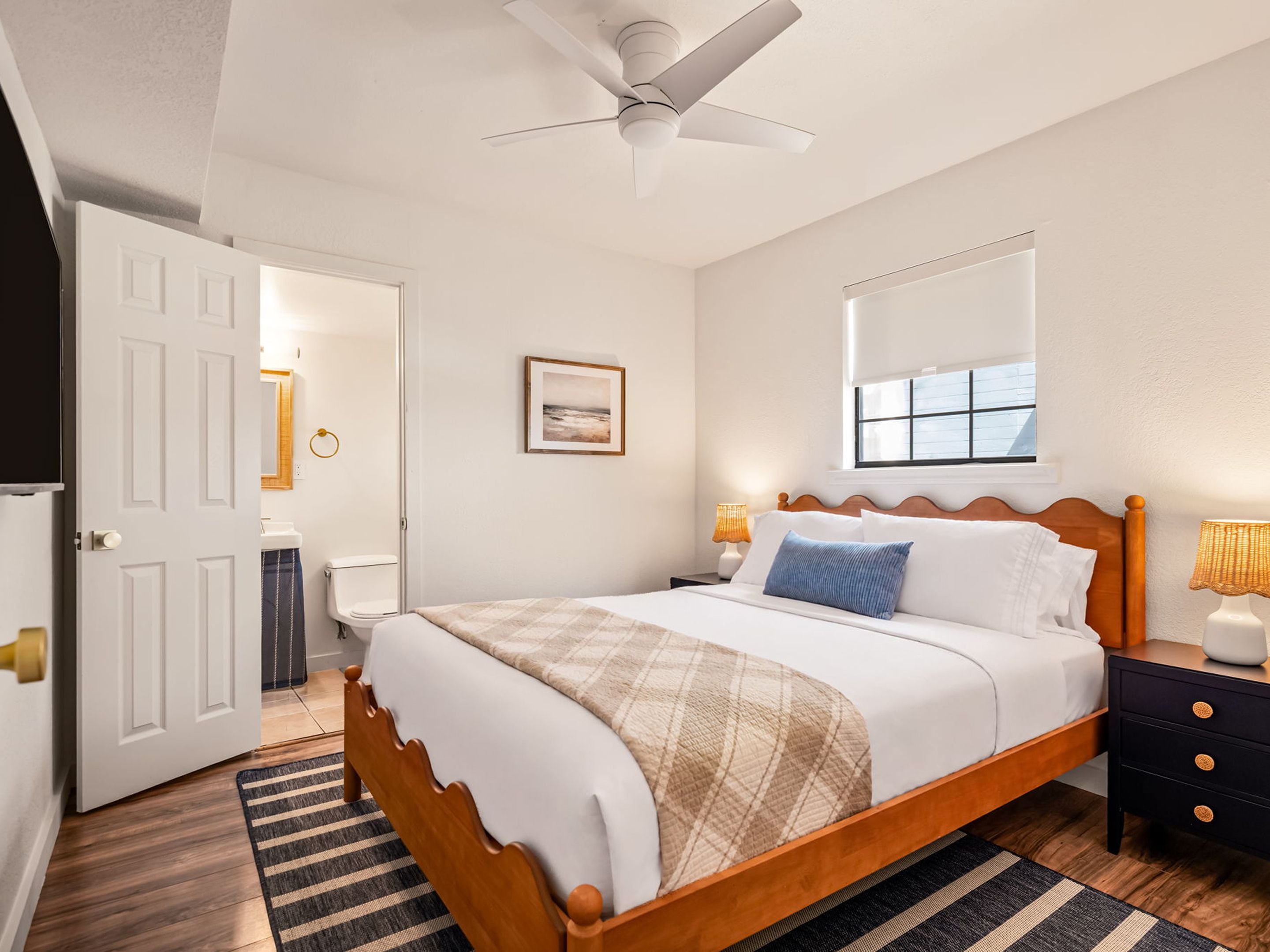 A bedroom with a wooden bed, a ceiling fan, a bedside table with a lamp, and a window with a view.