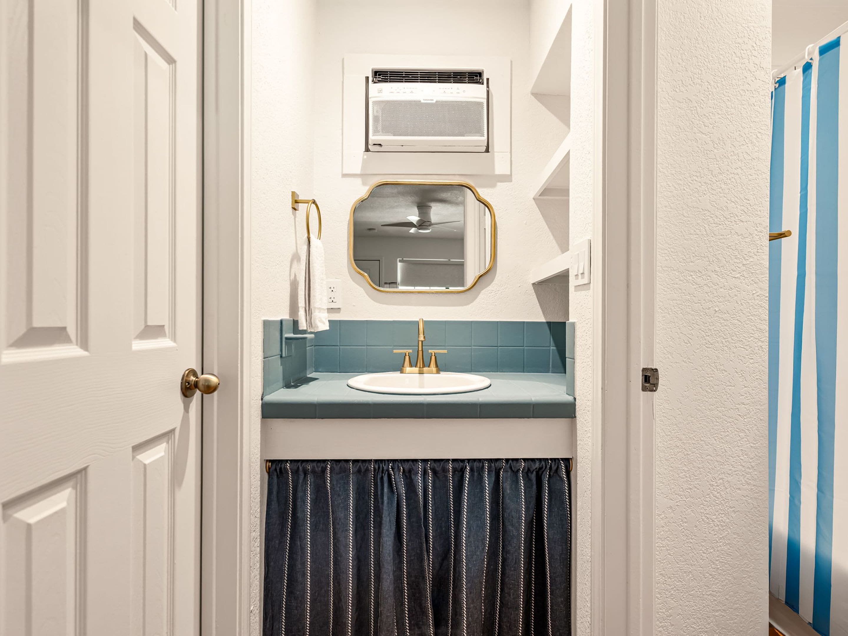 A bathroom with a white door, a white sink with a gold faucet, a mirror with a gold frame, and a towel hanging on the wall.