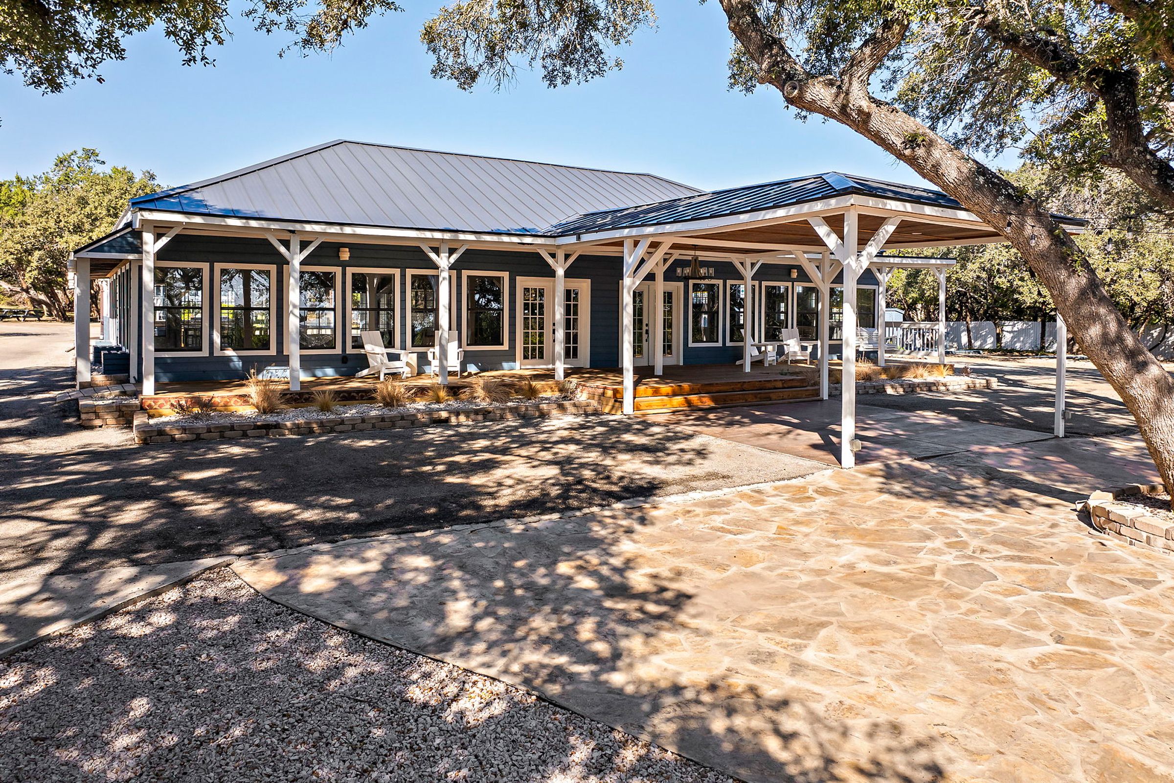 A covered outdoor seating area with multiple chairs and tables, a stone patio, and a large tree providing shade.