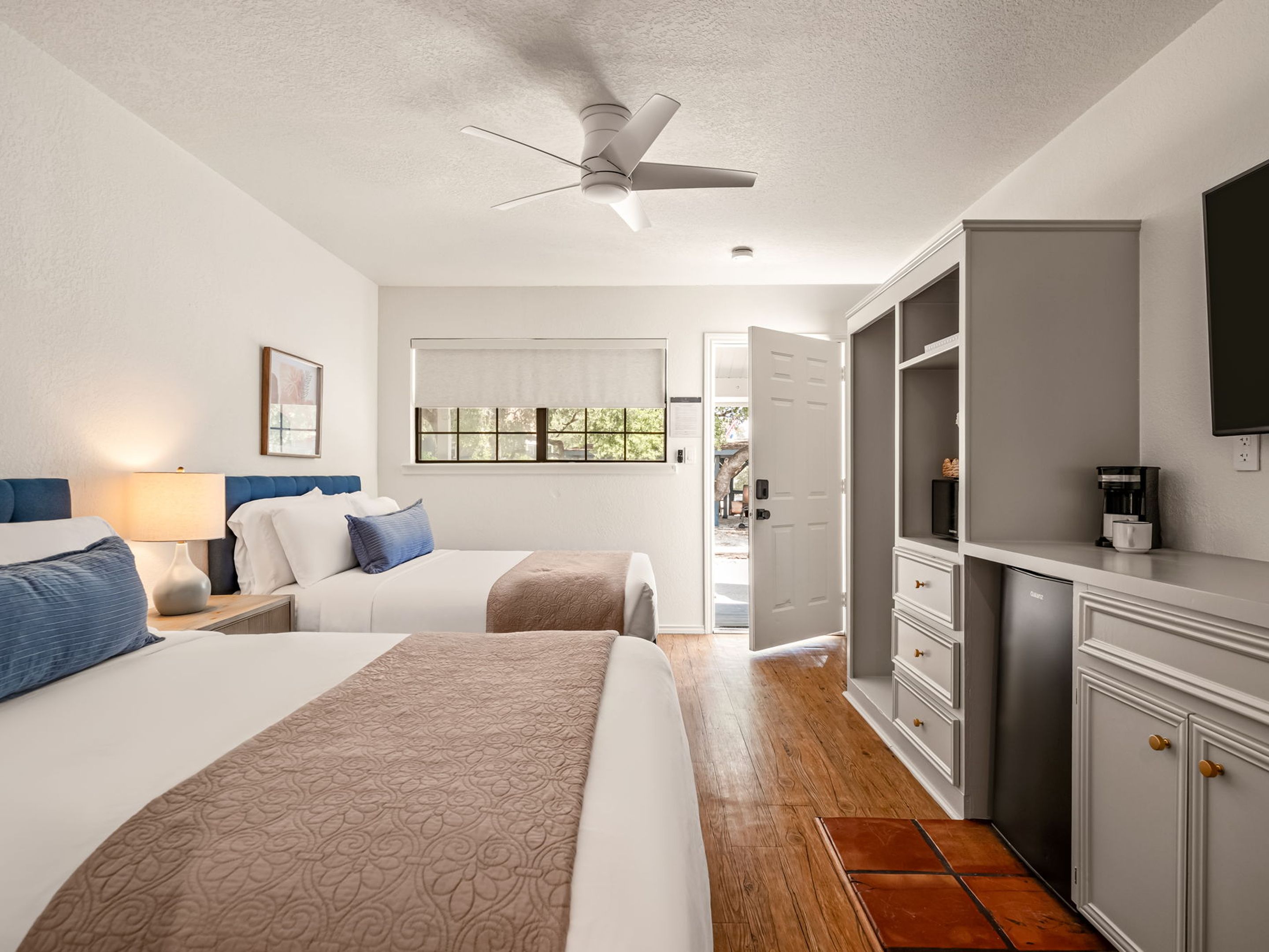 Habitación de hotel con dos camas, ventilador de techo, televisión, cafetera y vistas al exterior.