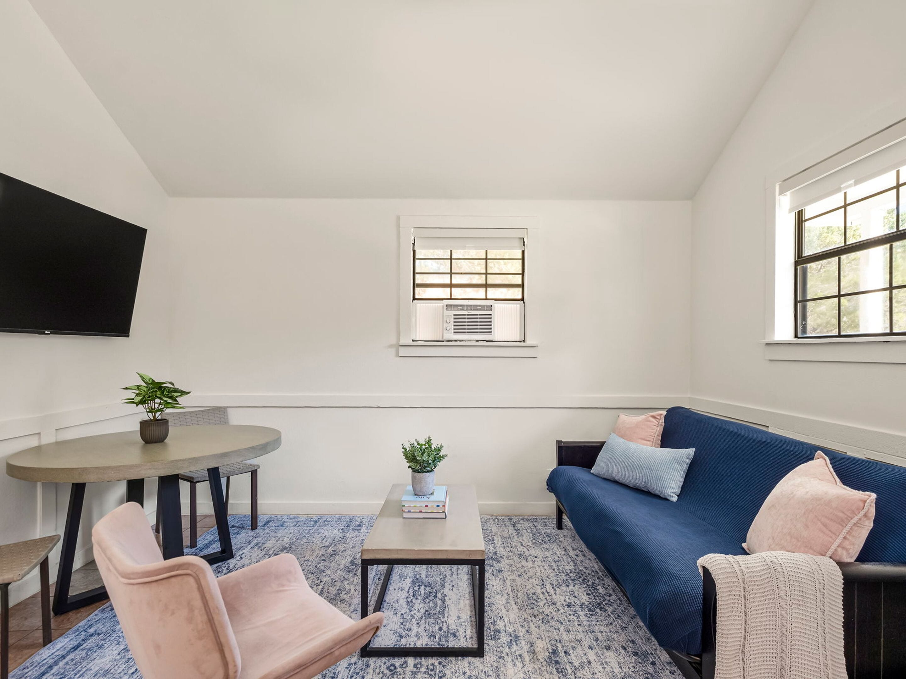 A living room with a blue sofa, a coffee table, a TV mounted on the wall, and a window with an air conditioner.