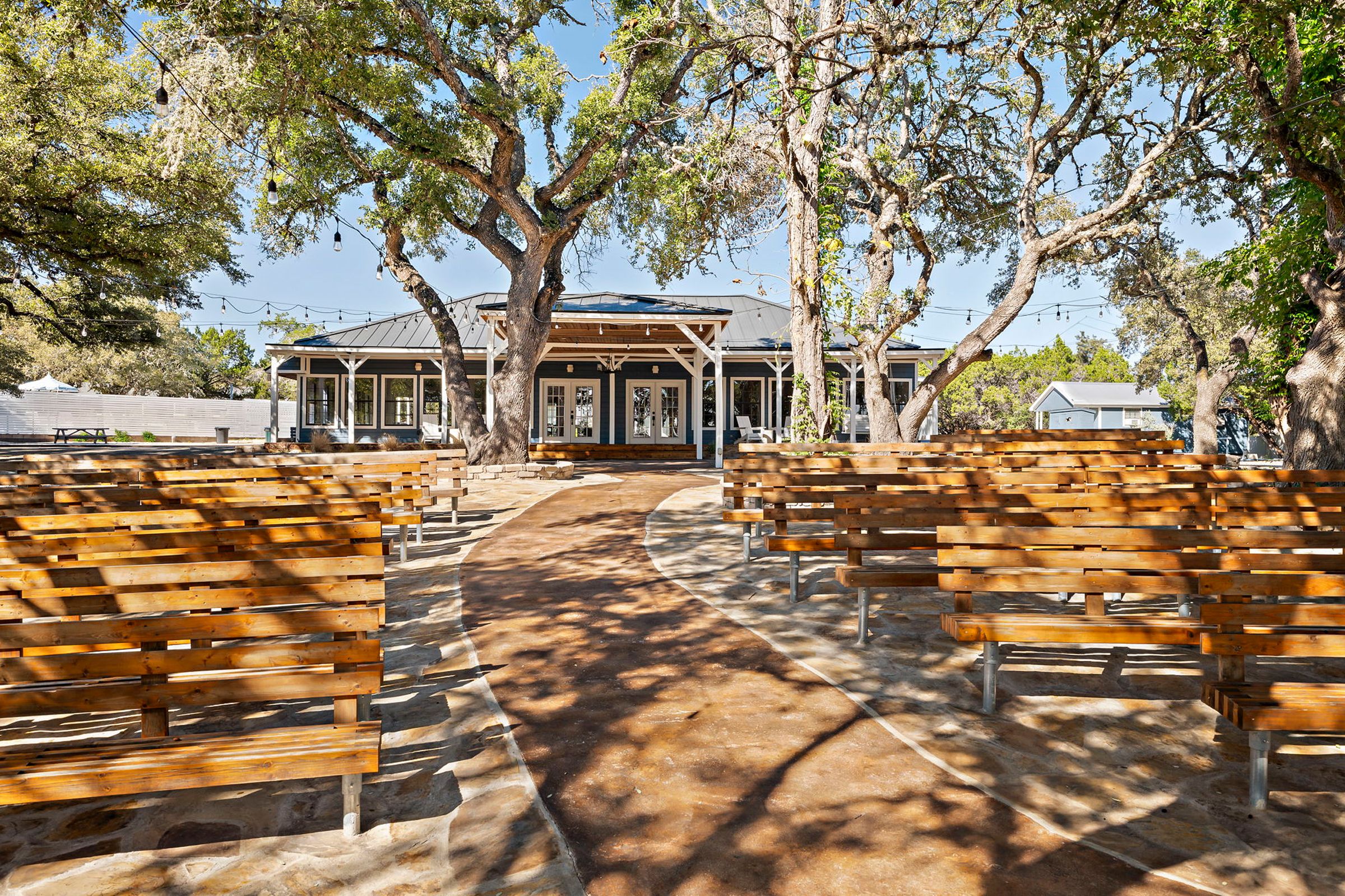 A covered outdoor area with wooden benches arranged in rows, a pathway leading to a building with a covered porch, and trees providing shade.