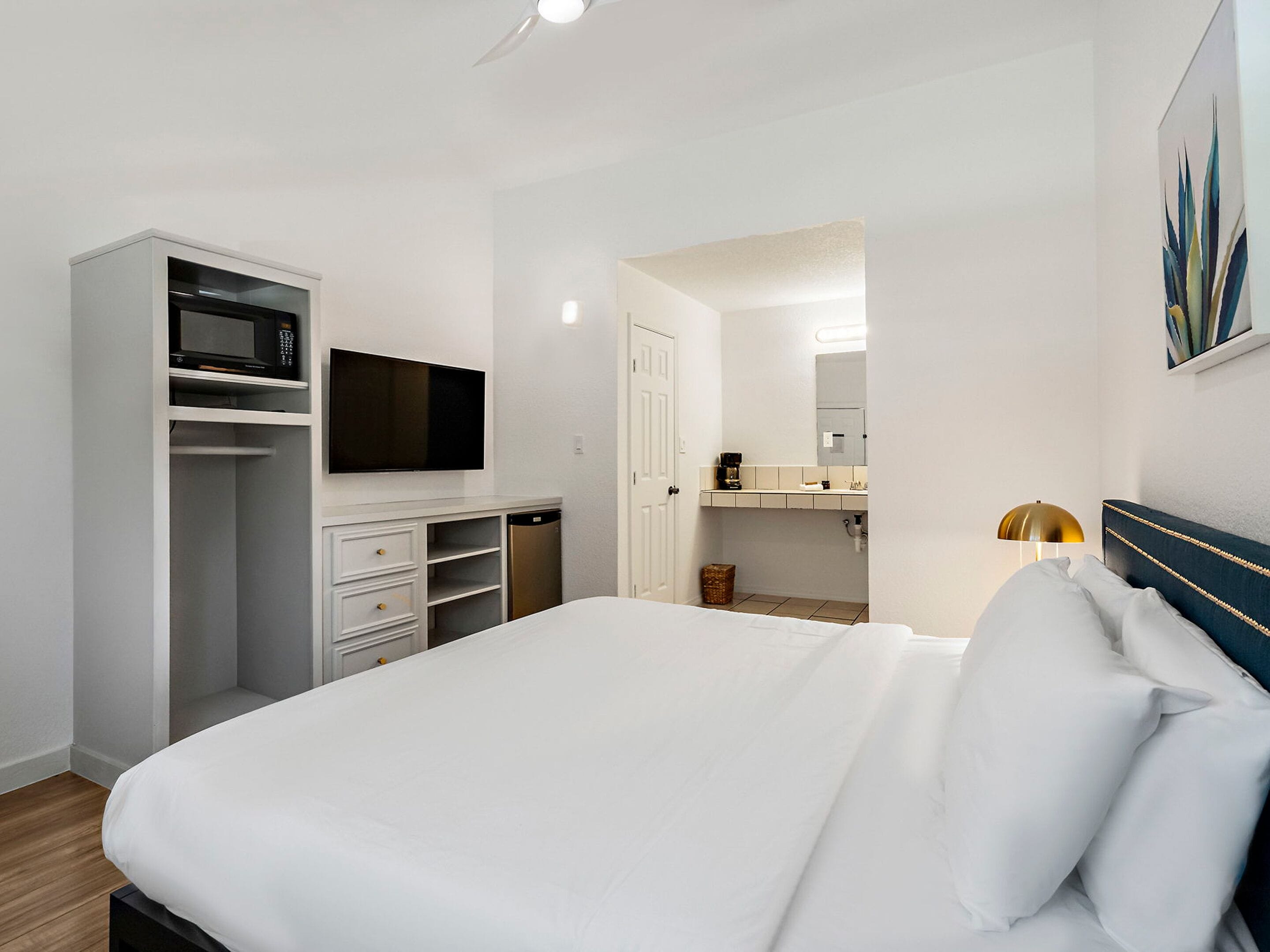 A modern hotel room with a bed, a TV, a wardrobe, a microwave, and a bathroom visible through an open door.