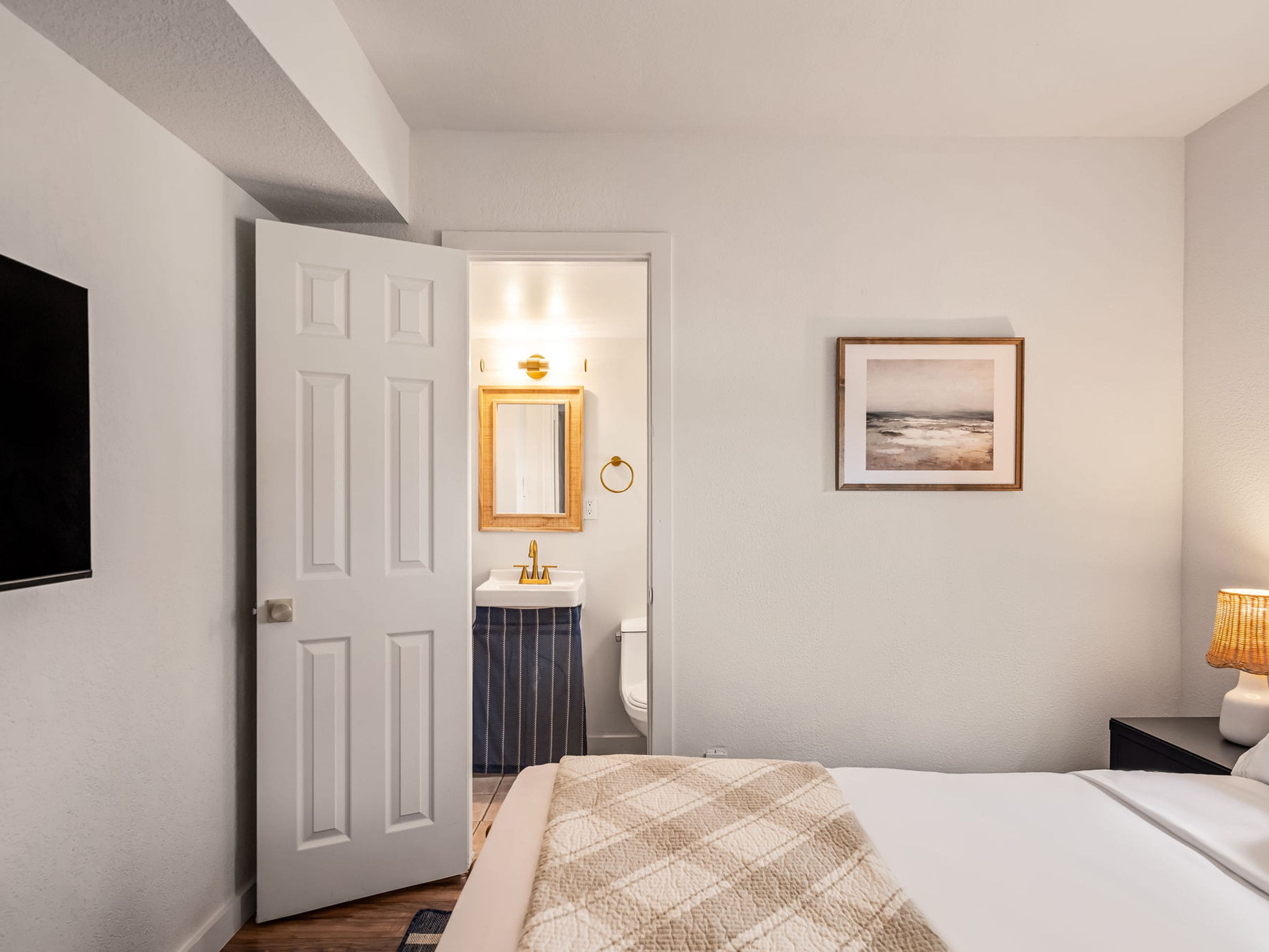 Quarto de hotel com cama, mesa de cabeceira, televisão presa à parede, quadro emoldurado e banheiro visível através de uma porta aberta.