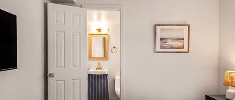 Une chambre d’hôtel avec un lit, une table de chevet, un téléviseur fixé au mur, un tableau encadré et une salle de bain visible à travers une porte ouverte.