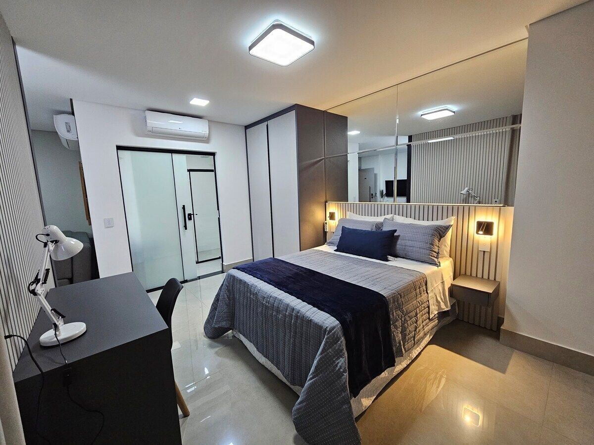Quarto moderno com cama, mesas de cabeceira, escrivaninha com cadeira e espelho.