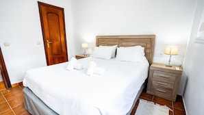 Una cama bien hecha con sábanas y almohadas blancas, un cabecero de madera, dos mesitas de noche con lámparas y un pequeño aparador.