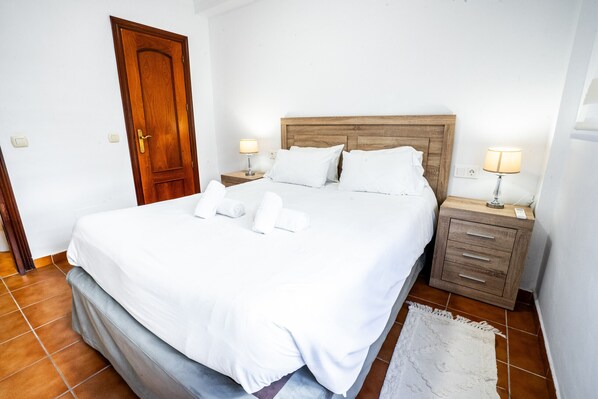 Una cama bien hecha con sábanas y almohadas blancas, un cabecero de madera, dos mesitas de noche con lámparas y un pequeño aparador.