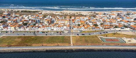 An aerial view of a coastal town with a beach, houses with orange roofs, and a tennis court.