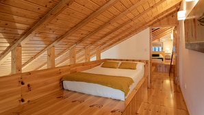 Un dormitorio con techo y suelo de madera, una cama con ropa blanca y un plaid color mostaza, y una mesita de noche con una lámpara.