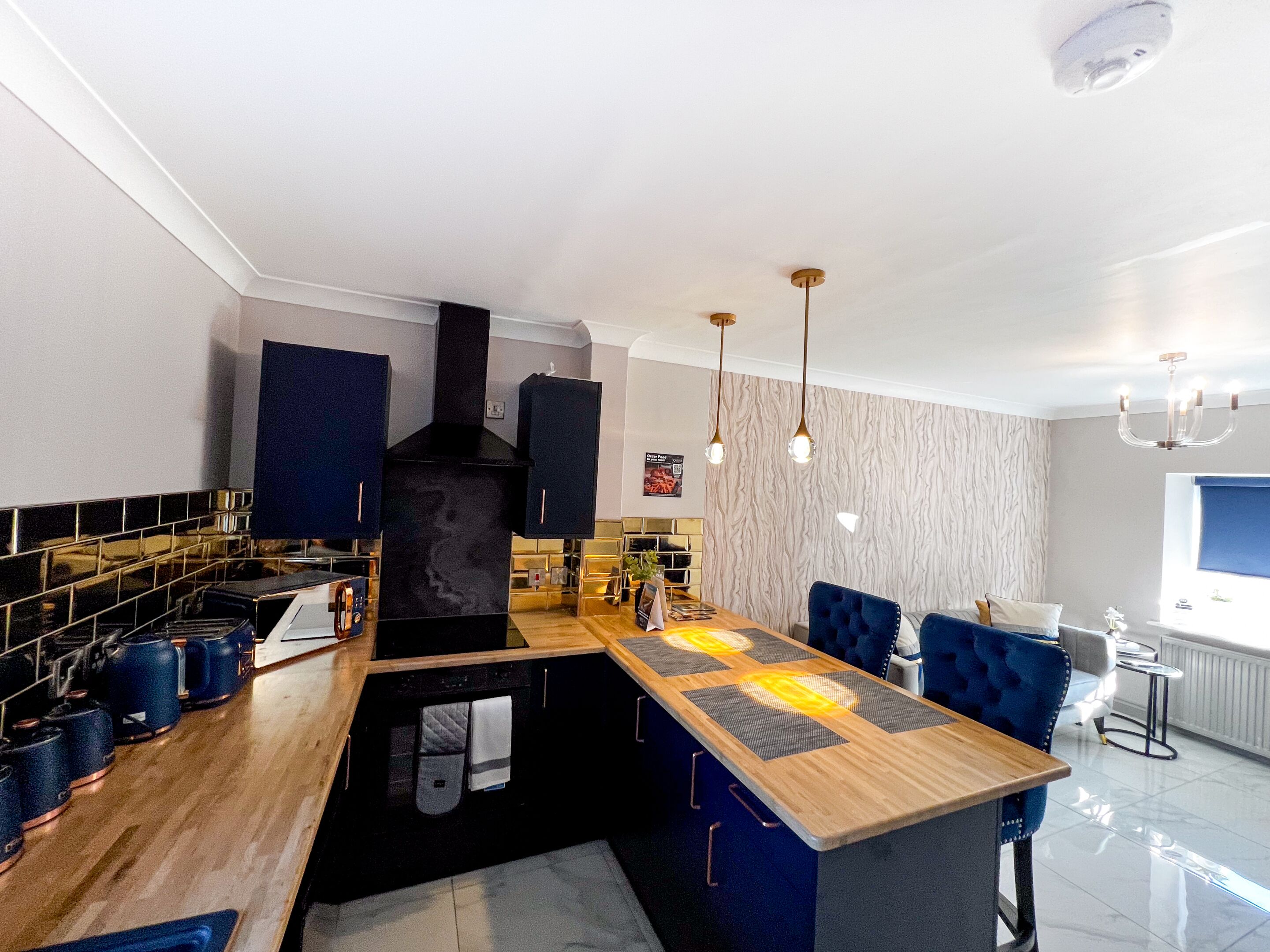 Una cocina moderna con una isla central, armarios negros y un horno integrado. Hay luces colgantes y una pequeña zona de comedor con sillas azules.