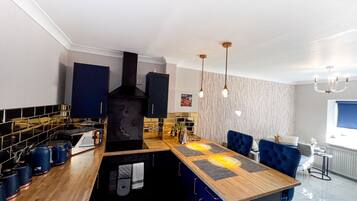 Una cocina moderna con una isla central, armarios negros y un horno integrado. Hay luces colgantes y una pequeña zona de comedor con sillas azules.