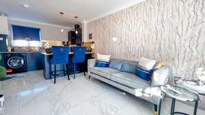 A modern living room with a grey sofa, blue chairs, a glass coffee table, and a kitchen area with a washing machine. - Lock View (Boston)