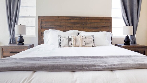 Una cama bien hecha con cabecera de madera, ropa de cama blanca y almohadones decorativos. Hay dos mesitas de noche con lámparas y una ventana con cortinas.