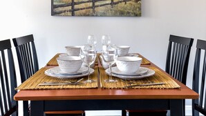 Una mesa de comedor dispuesta con platos, tazones y copas de vino blancos, rodeada de sillas negras.