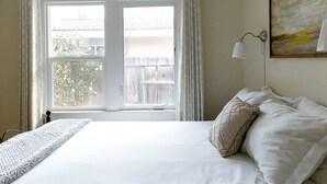 Un dormitorio con una cama grande, cortinas blancas, vista a una terraza de madera y un cuadro en la pared.